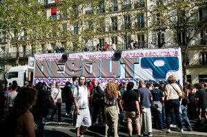 A crowd gathers on a city street in Paris in front of a large banner reading “DEBOUT” during a 2026 protest. Some people stand on a ledge, whilst trees and a block of flats are visible in the background, reminiscent of scenes from the annual Techno Parade.