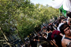 A large crowd gathers outdoors beneath green trees in Paris, with some people facing away from the camera. A white inflatable structure is visible on the right side of the image, suggesting a festive atmosphere—perhaps part of the 2026 Techno Parade.