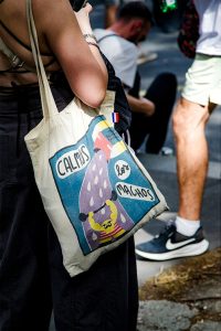 A person carries a tote bag with colourful artwork and the words “Calmos les machos,” whilst others sit or walk nearby in an outdoor setting during the lively Techno Parade in Paris, 2026.
