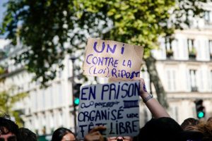 People at a protest hold handwritten signs in French, one reading “UNi CONTRE LA [LOI] RIPOSTE” and another mentioning “EN PRISON” and “ORGANISÉ UNE TÊT LIBRE,” with buildings and trees in the background—a vivid scene reminiscent of the spirited gatherings seen during events like the Techno Parade in Paris.