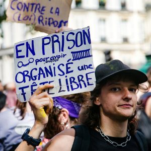 A person at a protest in Paris holds a handwritten sign in French that reads, “En prison pour avoir organisé une tête libre,” surrounded by other protesters and blurred buildings in the background. The scene captures the spirit of activism during the 2026 Techno Parade.