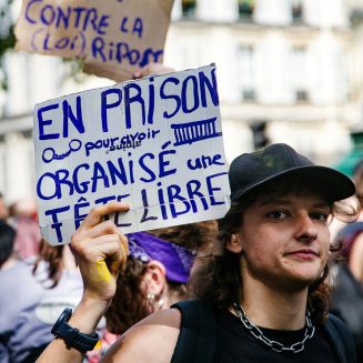 20260919-5H1A0261_MOD_FRAME_resize A person at a protest in Paris holds a handwritten sign in French that reads, “En prison pour avoir organisé une tête libre,” surrounded by other protesters and blurred buildings in the background. The scene captures the spirit of activism during the 2026 Techno Parade.