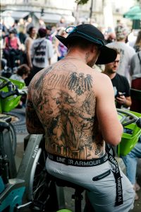 A man with an elaborate back tattoo sits on a public bicycle in a crowded outdoor setting, wearing a black hat and Under Armour shorts. The vibrant atmosphere hints at the excitement of the Paris 2026 Techno Parade, with other people and bicycles visible in the background.