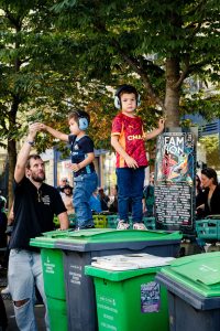 Two young boys wearing ear defenders stand on recycling bins at an outdoor event, with an adult nearby. One boy is in a red shirt, the other in blue. Trees and a colourful Techno Parade 2026 poster hint at the lively atmosphere in Paris, visible in the background.