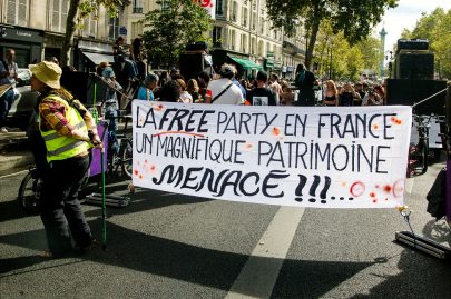 20260919-5H1A0267_MOD_FRAME_resize Amidst the vibrant atmosphere of Paris, a group holds a large banner at a street protest, reading “La free party en France, un magnifique patrimoine menacé!!!” People and trees line the background, capturing the spirited scene reminiscent of the city’s famed Techno Parade as France looks ahead to 2026.