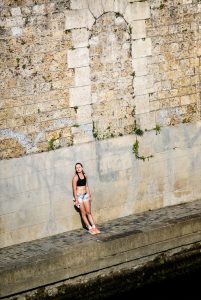 A woman in shorts and a black top stands against a stone wall with overgrown ivy, holding a camera and looking up, bathed in sunlight.