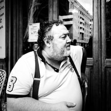 Jean Benoit Roubinet, a man with a beard wearing a polo shirt and braces, sits outside a shop window in Paris.