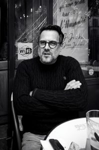 A man with glasses and a beard, Jean Benoit Roubinet, sits with his arms crossed at a café table in front of a window with cracked glass and French text.