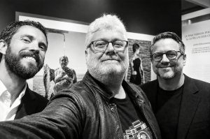 Three men pose for a selfie at the Salon de la photo in Paris, surrounded by an exhibition of black-and-white photography.