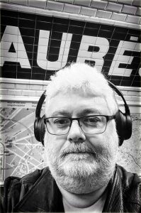 A selfie of Jean Benoit Roubinet, a bearded man wearing glasses and headphones, standing in front of an AUBER metro sign and underground map while attending the Salon de la photo in Paris.
