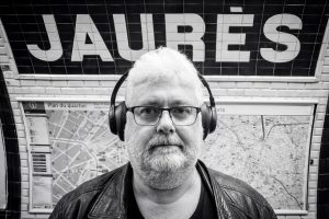 Jean Benoit Roubinet wears headphones and glasses whilst standing in front of a Jaurès Paris Underground station sign and a neighbourhood map.