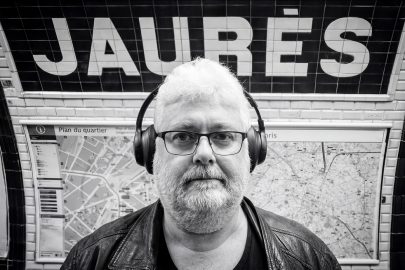 Jean Benoit Roubinet wears headphones and glasses whilst standing in front of a Jaurès Paris Underground station sign and a neighbourhood map.