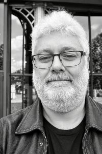 A black and white portrait of Jean Benoit Roubinet, a man with a grey beard and glasses wearing a leather jacket.