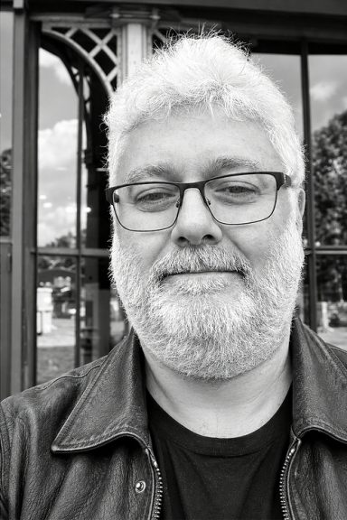 A black and white portrait of Jean Benoit Roubinet, a man with a grey beard and glasses wearing a leather jacket.